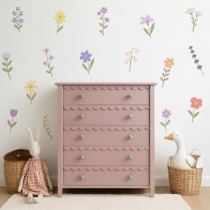 Peut inclure: Une commode rose aux bords festonn&eacute;s et aux boutons verts se dresse contre un mur blanc orn&eacute; de d&eacute;calcomanies florales color&eacute;es. Un lapin en peluche et un canard dans un panier compl&egrave;tent le th&egrave;me de la chambre d'enfant.