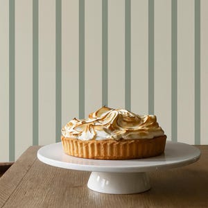 May include: A golden-brown tart with a meringue topping sits on a white cake stand. The dessert is on a wooden table, with a striped background of light beige and muted green vertical lines.