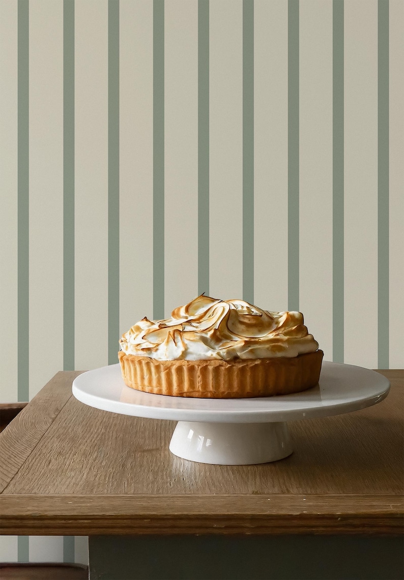May include: A golden-brown tart with a meringue topping sits on a white cake stand. The dessert is on a wooden table, with a striped background of light beige and muted green vertical lines.