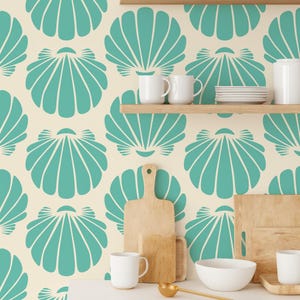 May include: A kitchen scene with a cream-colored wall featuring a repeating teal seashell pattern. Wooden shelves hold white mugs, plates, and a teapot. A white countertop displays cutting boards, a bowl, and a gold ladle.