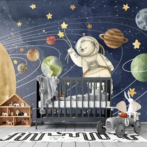 May include: A nursery scene with a dark gray crib, a toy car, and a dollhouse. The wall features a watercolor mural of planets, stars, and a bear astronaut. A gray blanket with pom-poms is draped over the crib.