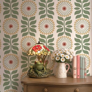 May include: A table lamp featuring a green frog base and a stained glass shade with red flowers, positioned next to a white pitcher filled with white daisies and a row of four books, all set against wallpaper with a repeating green and red floral pattern.
