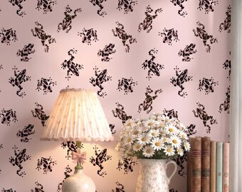 May include: A light pink wallpaper featuring a repeating pattern of dark brown frogs. In front of the wall sits a table lamp with a pleated shade and a bow detail, a white pitcher filled with white daisies, and a row of four vintage books.