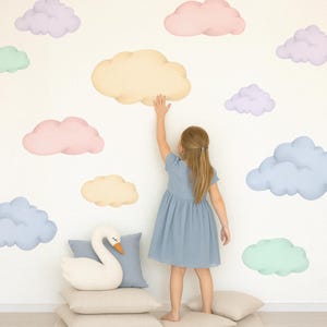 May include: A young person in a blue dress reaches towards a cloud-shaped wall decal. The wall is decorated with various pastel-colored cloud decals. A white swan-shaped pillow rests on a stack of beige pillows, creating a whimsical nursery theme.