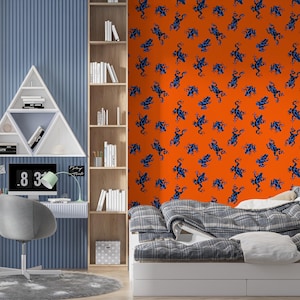 Op de afbeelding: Een moderne slaapkamer met een blauw en oranje kleurenschema. De oranje muur heeft een patroon van blauwe kikkers. Een wit bureau met een computer en een grijze stoel staan voor een blauwe muur. Een witte boekenkast staat rechts.