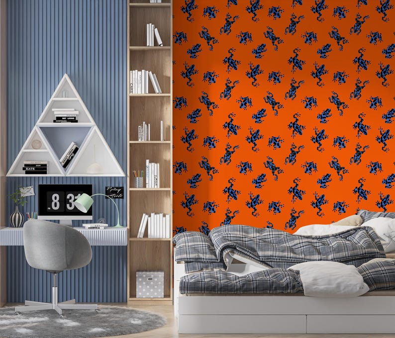 Op de afbeelding: Een moderne slaapkamer met een blauw en oranje kleurenschema. De oranje muur heeft een patroon van blauwe kikkers. Een wit bureau met een computer en een grijze stoel staan voor een blauwe muur. Een witte boekenkast staat rechts.