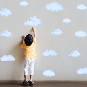 May include: A child in a yellow shirt and white pants reaching towards a light blue cloud decal on a beige wall. Several other cloud decals are scattered around the wall, creating a whimsical scene.