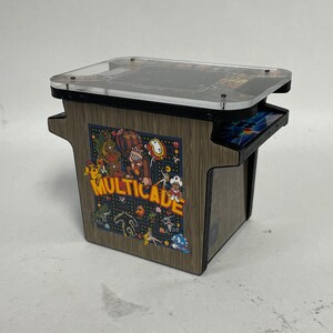 May include: A miniature arcade cabinet with a wood grain finish. The cabinet features a clear plastic top and a colorful graphic of various video game characters. The text "MULTICADE" is printed on the front of the cabinet.