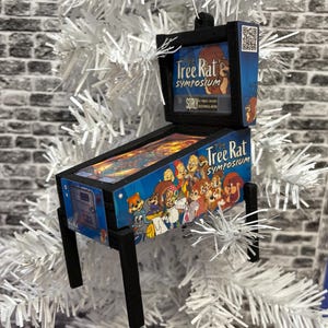 May include: A miniature pinball machine ornament with the text "Tree Rat Symposium" on a blue background. The ornament features cartoon characters and is supported by black legs. It is displayed on a white Christmas tree.