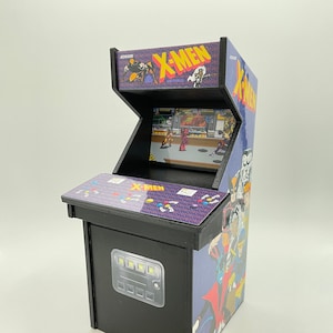 May include: A miniature arcade cabinet with a black wooden base and a purple and black front panel. The front panel features a graphic of the X-Men characters. The cabinet has a small screen and a control panel with buttons and a joystick. The cabinet is a replica of the classic X-Men arcade game.