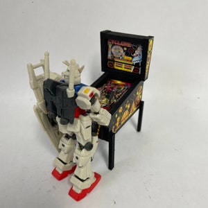 May include: A white and gray robot figure stands next to a black pinball machine with a yellow and red graphic on the front. The pinball machine has the text "CYCLONE" on the front.