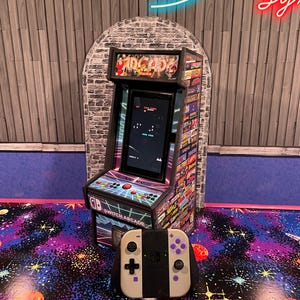 May include: A miniature Switch Arcade cabinet with a black screen displaying a game. The cabinet is decorated with colorful arcade game graphics and the words "Switch Arcade". A white and purple controller sits in front of the cabinet.
