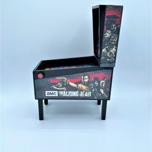 Puede incluir: Una m&aacute;quina de pinball en miniatura de color negro y rojo con un gr&aacute;fico de "The Walking Dead" que presenta a Rick Grimes y otros personajes. La m&aacute;quina tiene un bot&oacute;n rojo y un campo de juego transparente.