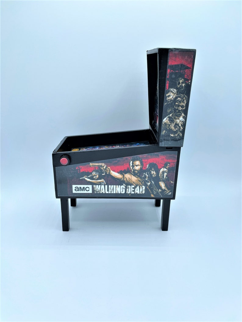 Puede incluir: Una m&aacute;quina de pinball en miniatura de color negro y rojo con un gr&aacute;fico de "The Walking Dead" que presenta a Rick Grimes y otros personajes. La m&aacute;quina tiene un bot&oacute;n rojo y un campo de juego transparente.