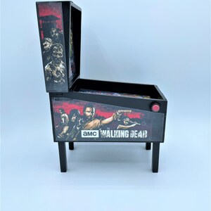 Puede incluir: Una m&aacute;quina de pinball en miniatura de color negro y rojo con el tema de "The Walking Dead". La m&aacute;quina presenta un gr&aacute;fico de personajes del programa, incluidos Rick Grimes, Daryl Dixon y Michonne. La m&aacute;quina tiene un bot&oacute;n rojo en el lateral y el texto "AMC The Walking Dead" en la parte delantera.