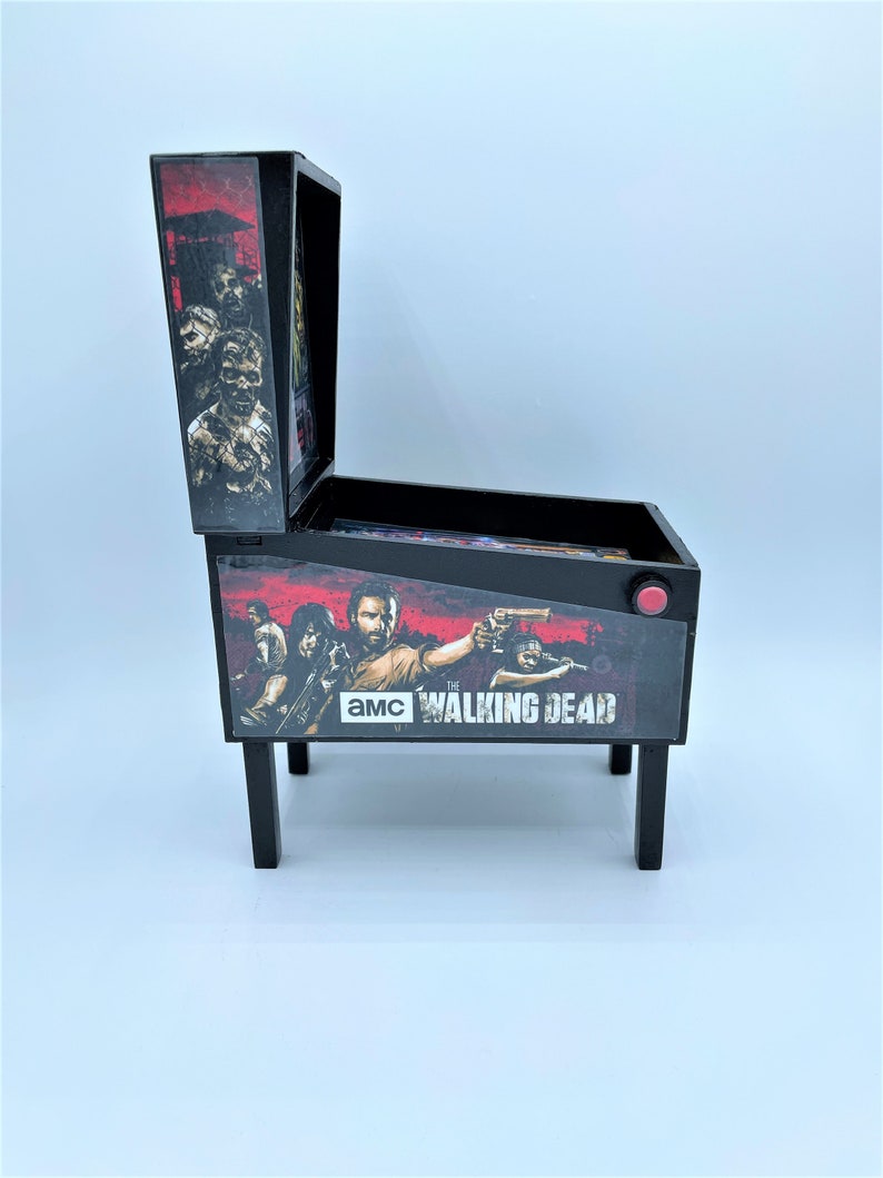 Puede incluir: Una m&aacute;quina de pinball en miniatura de color negro y rojo con el tema de "The Walking Dead". La m&aacute;quina presenta un gr&aacute;fico de personajes del programa, incluidos Rick Grimes, Daryl Dixon y Michonne. La m&aacute;quina tiene un bot&oacute;n rojo en el lateral y el texto "AMC The Walking Dead" en la parte delantera.
