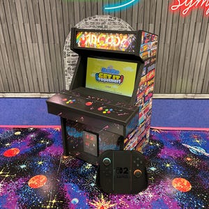 May include: A black arcade machine with colorful game art and the word "ARCADE" on top. The screen displays a game title. A black Nintendo Switch controller sits in front of the machine on a galaxy-themed rug. The words "Home of Squirrel" are visible.