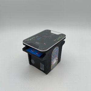 May include: A black and green miniature arcade cabinet with a clear plastic top. The cabinet has a blue screen with the words "HOME" and "FIRE" on it. The cabinet has a sticker on the side with the words "Galaga" and a picture of a spaceship.