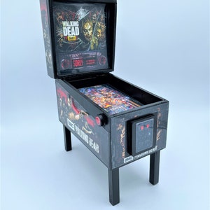 Puede incluir: Una m&aacute;quina de pinball negra en miniatura con el tema de "The Walking Dead". La m&aacute;quina tiene un frente de vidrio con un gr&aacute;fico de personajes del programa. La m&aacute;quina est&aacute; sobre un fondo blanco.