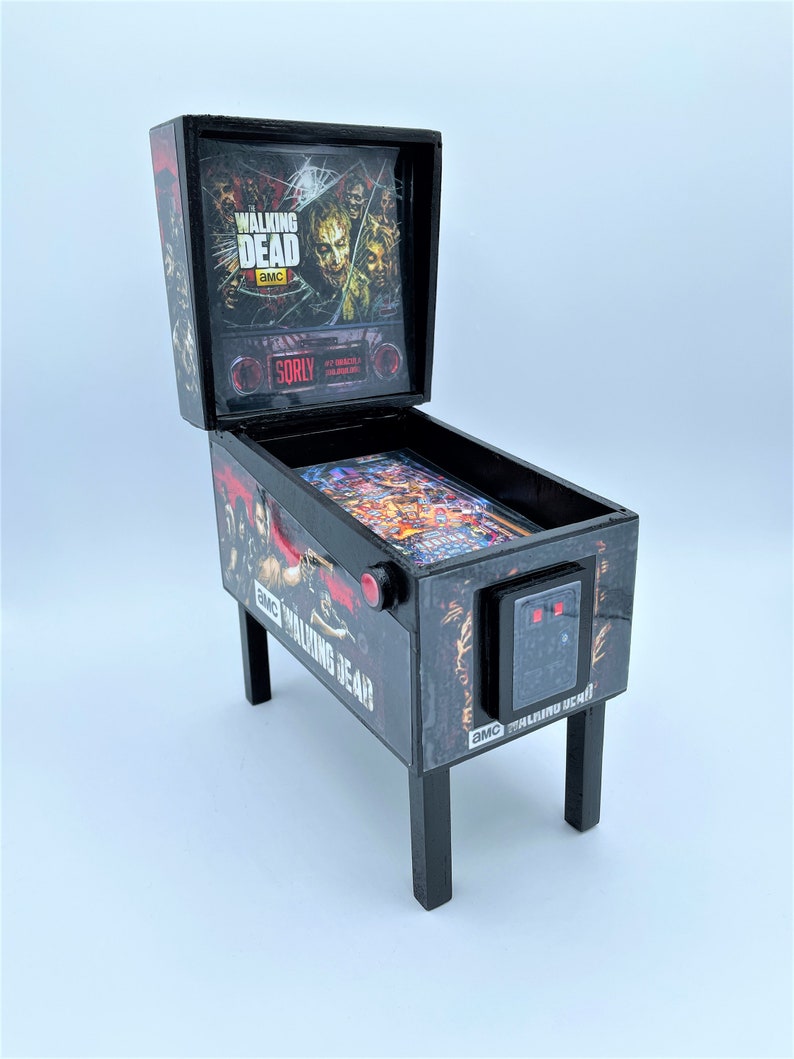 Puede incluir: Una m&aacute;quina de pinball negra en miniatura con el tema de "The Walking Dead". La m&aacute;quina tiene un frente de vidrio con un gr&aacute;fico de personajes del programa. La m&aacute;quina est&aacute; sobre un fondo blanco.