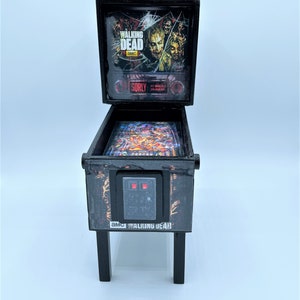 Puede incluir: Una m&aacute;quina de pinball en miniatura de color negro con el tema de "The Walking Dead". La m&aacute;quina tiene un frente de vidrio con un gr&aacute;fico de zombis del programa. La m&aacute;quina tiene un gabinete negro con un letrero rojo "SORRY" y un gr&aacute;fico en blanco y negro de una mano de zombi. La m&aacute;quina tiene una base negra con cuatro patas.