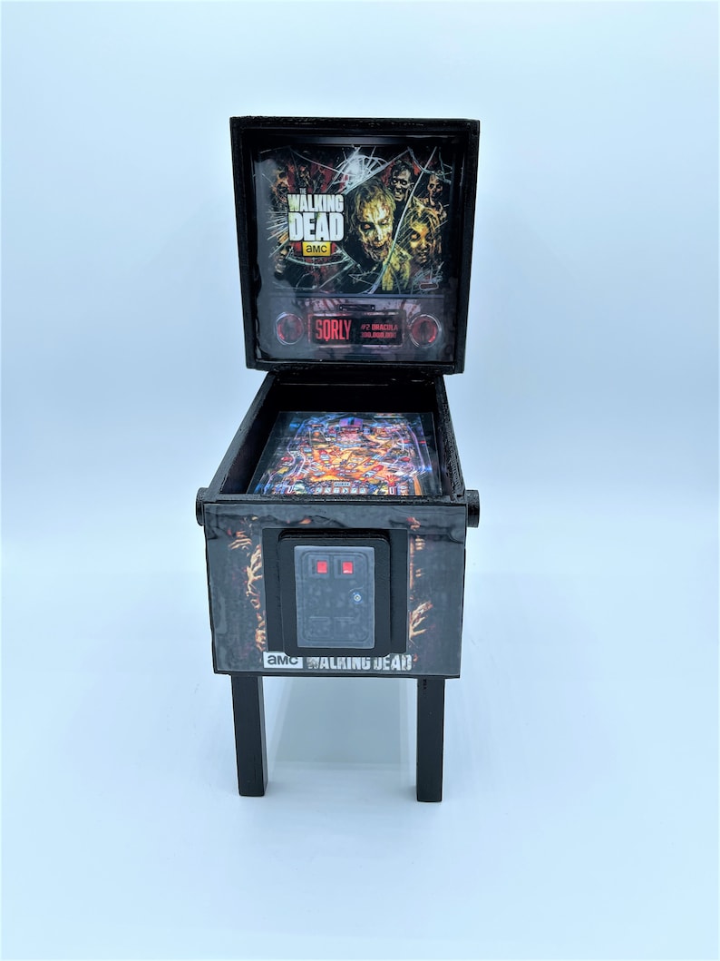 Puede incluir: Una m&aacute;quina de pinball en miniatura de color negro con el tema de "The Walking Dead". La m&aacute;quina tiene un frente de vidrio con un gr&aacute;fico de zombis del programa. La m&aacute;quina tiene un gabinete negro con un letrero rojo "SORRY" y un gr&aacute;fico en blanco y negro de una mano de zombi. La m&aacute;quina tiene una base negra con cuatro patas.