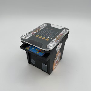 May include: A miniature arcade cabinet with a clear plastic top. The cabinet is black with a yellow and orange graphic of a chef. The game screen shows a pixelated game with the text "BurgerTime" on the top and bottom.
