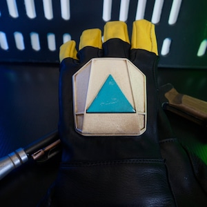May include: A black leather glove with a yellow fingertip design and a large, silver-colored, triangular emblem with a blue center.