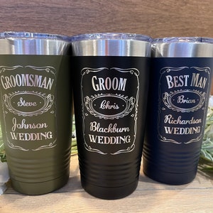 May include: Three stainless steel tumblers with personalized engravings for a wedding party. The tumblers are green, black, and blue. The text on the tumblers reads "Groom", "Best Man", and "Groomsman" with the names "Chris", "Brian", and "Steve" respectively. The text also includes the last names and the word "Wedding".