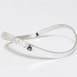 May include: A silver bangle bracelet with a hammered texture. The bracelet has a flowing, open design, with a slightly flattened band. The metal has a bright, reflective quality, and appears to be made of sterling silver.