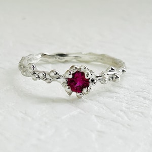 May include: A silver ring with a floral design. The ring features a round, vibrant red gemstone at the center, surrounded by a detailed, textured setting. The band has a delicate, organic pattern, resembling leaves and vines.