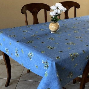 May include: A blue tablecloth with olive branch patterns on a wooden table. The tablecloth has a light blue background with green and black olive branch designs. Two wooden chairs are behind the table, and a vase of white flowers sits on the table.