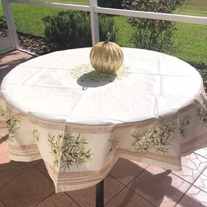 Puede incluir: Un mantel redondo blanco con un patr&oacute;n de ramas de olivo verdes y una calabaza decorativa dorada en el centro.