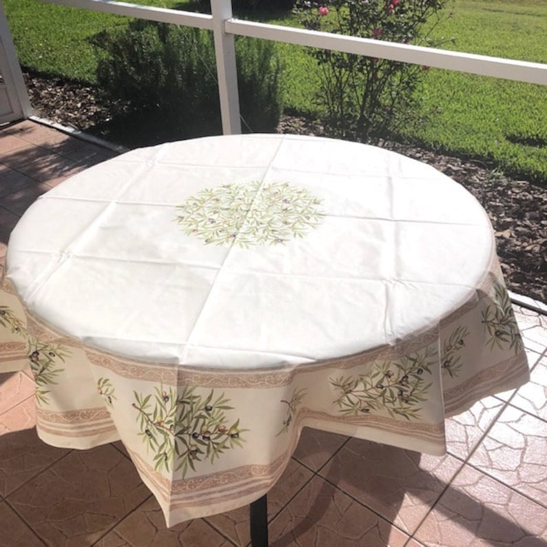 Puede incluir: Un mantel redondo blanco con un dise&ntilde;o de rama de olivo verde. El mantel tiene un borde marr&oacute;n con un patr&oacute;n repetitivo. El mantel est&aacute; sobre una mesa con una base de metal blanco.