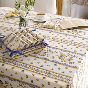 May include: A white tablecloth with blue and yellow stripes and a floral pattern featuring birds. The tablecloth is set with a vase of white flowers, a cup and saucer, a book, and a stack of folded napkins.