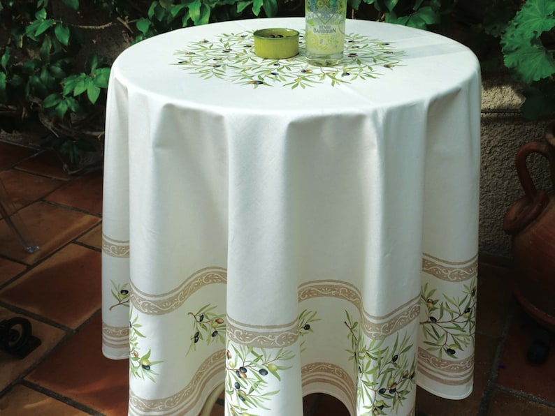 Puede incluir: Mantel redondo blanco con un dise&ntilde;o de rama de olivo verde y un borde beige. El mantel est&aacute; sobre una mesa redonda con un vaso de agua y un plato verde en la parte superior.