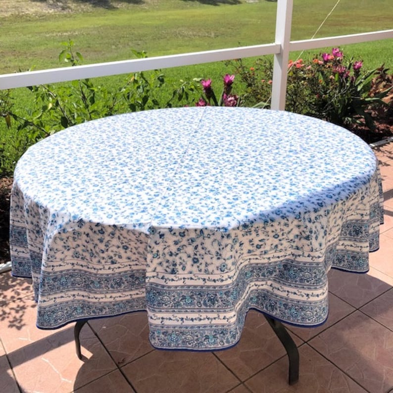 May include: A round tablecloth with a white background and a blue floral pattern. The border features a blue and white floral design. The tablecloth is draped over a dark metal table.