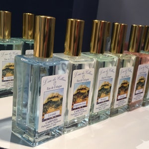 K&ouml;nnte beinhalten: Eine Sammlung rechteckiger Glas-Cologne-Flaschen mit goldfarbenen Verschl&uuml;ssen. Jede Flasche hat ein Etikett mit einer Landschaftsillustration und Text. Die Flaschen sind mit unterschiedlich farbigen Fl&uuml;ssigkeiten gef&uuml;llt, darunter Blau, Gr&uuml;n und Rosa.