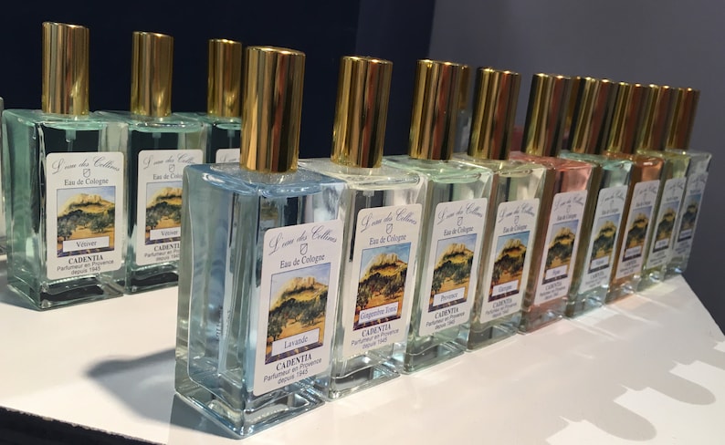 K&ouml;nnte beinhalten: Eine Sammlung rechteckiger Glas-Cologne-Flaschen mit goldfarbenen Verschl&uuml;ssen. Jede Flasche hat ein Etikett mit einer Landschaftsillustration und Text. Die Flaschen sind mit unterschiedlich farbigen Fl&uuml;ssigkeiten gef&uuml;llt, darunter Blau, Gr&uuml;n und Rosa.