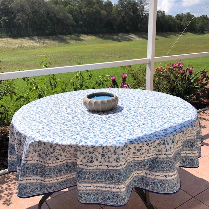 May include: A round tablecloth with a blue floral pattern and a decorative border. A small, gray bowl with a blue candle sits in the center of the table. The table is on a patio overlooking a green landscape.