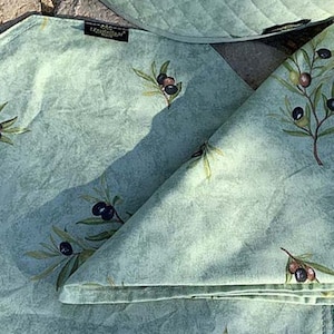 May include: A green fabric napkin with a pattern of olive branches and black olives. The napkin is folded in half and has a small tag with the word "Treasures" on it.