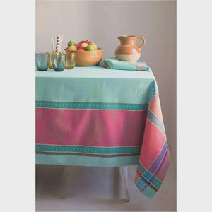 May include: A turquoise tablecloth with a pink and turquoise patterned border. A bowl of fruit, glasses, a pitcher, and a napkin are on the table. The tablecloth has a decorative design and is draped over a table.