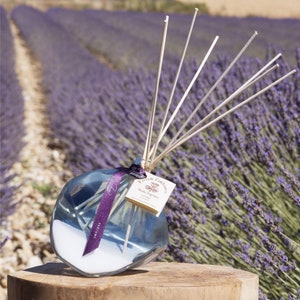 May include: A clear glass diffuser bottle with a purple ribbon and a label that reads "Parfum d'ambiance Home Fragrance Lavender". The bottle is filled with a clear liquid and has wooden sticks sticking out of it. The bottle is sitting on a wooden surface in front of a field of lavender.