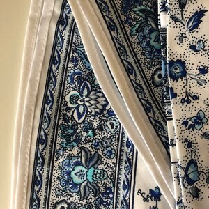 May include: Close-up of fabric with a white background and blue floral patterns. The fabric has a detailed design with various shades of blue, including teal and navy. The patterns include flowers and other decorative elements.