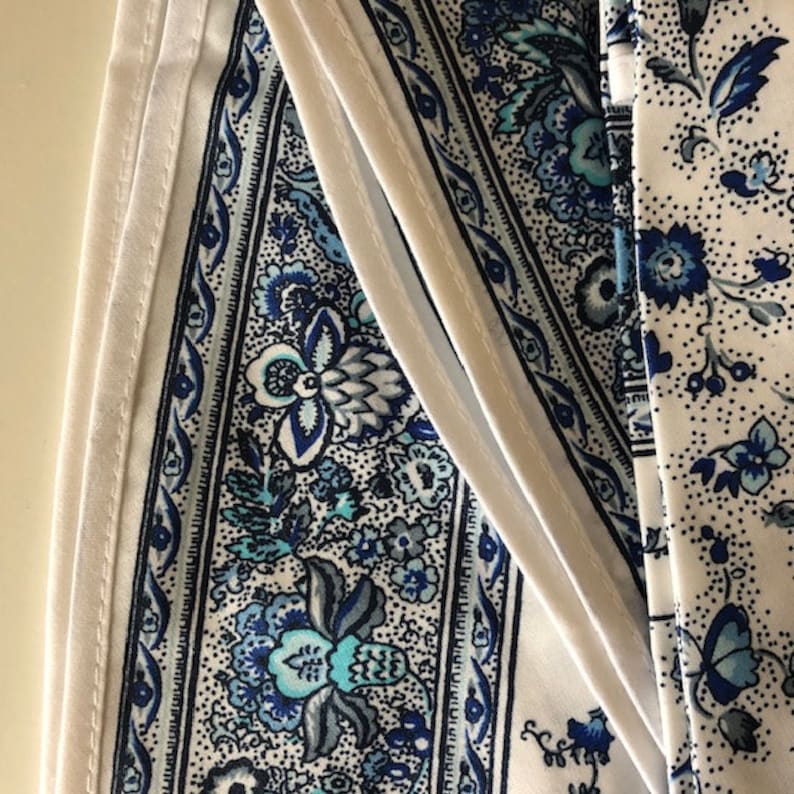 May include: Close-up of fabric with a white background and blue floral patterns. The fabric has a detailed design with various shades of blue, including teal and navy. The patterns include flowers and other decorative elements.