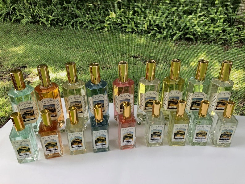K&ouml;nnte beinhalten: Eine Auswahl an Glasparf&uuml;mflaschen mit goldfarbenen Verschl&uuml;ssen. Die Flaschen enthalten Fl&uuml;ssigkeiten in verschiedenen Farben, darunter Gr&uuml;n, Gelb und Rosa. Jede Flasche hat ein Etikett mit Text und einem Landschaftsbild. Die Flaschen sind auf einer wei&szlig;en Oberfl&auml;che angeordnet.