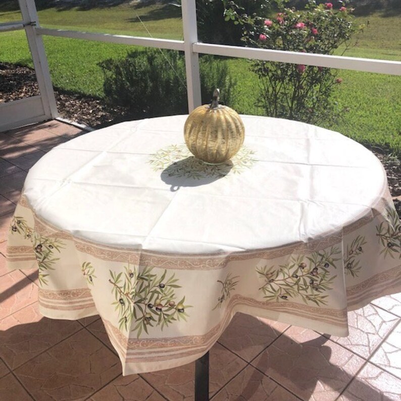 Puede incluir: Un mantel redondo blanco con un patr&oacute;n de ramas de olivo verdes y una calabaza decorativa dorada en el centro.