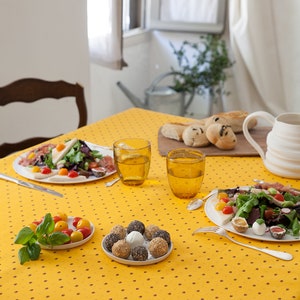Puede incluir: Un mantel amarillo con un pequeño patrón de lunares marrones. Una mesa puesta con dos vasos de agua, un plato de ensalada, un plato de pequeños bocadillos redondos y un plato de tomates cherry.
