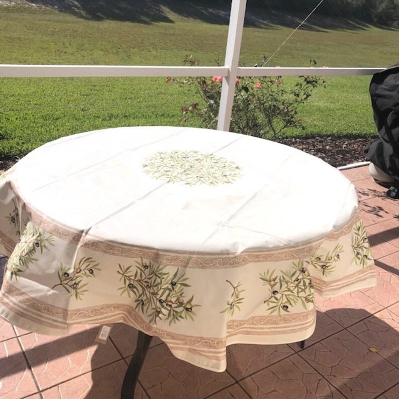 Puede incluir: Mantel redondo blanco con un dise&ntilde;o de ramas de olivo verdes. El mantel tiene un borde con un patr&oacute;n repetido de ramas de olivo y un fondo marr&oacute;n claro.