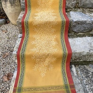 May include: A yellow table runner with a white olive branch design and a red, green, and brown border. The runner is on a set of stone steps.